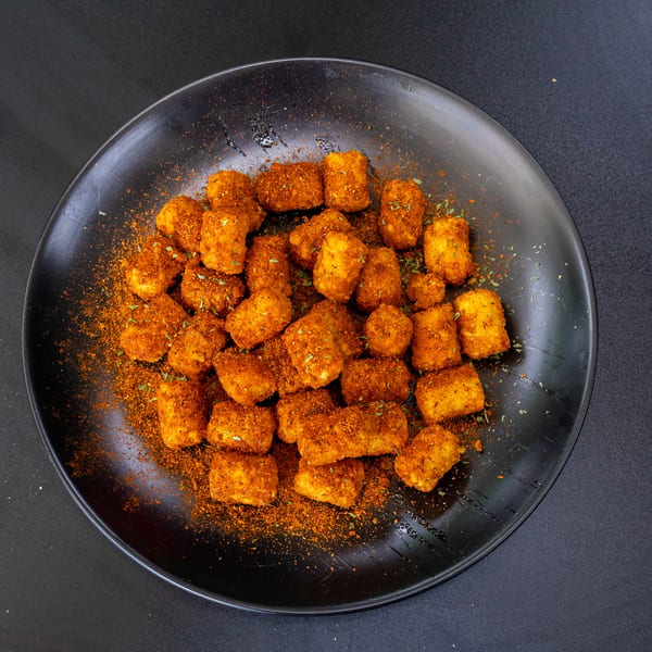 A black plate filled with golden-brown tater tots, generously seasoned with red spices and green herbs.