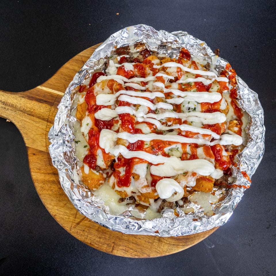 Loaded tater tots with cheese, ground meat, ketchup, and white sauce in foil on a wooden board.