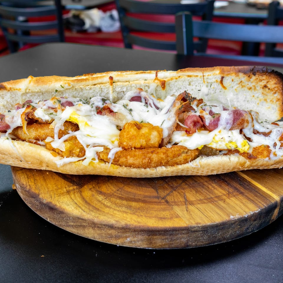 A large breakfast sub sandwich with fried chicken, tater tots, bacon, egg, and melted cheese.