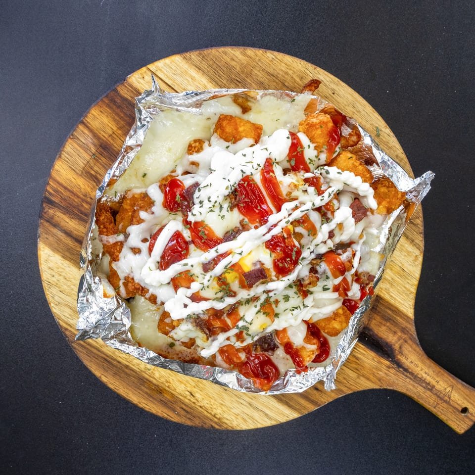 Loaded tater tots with melted cheese, ketchup, mayonnaise, and herbs on a wooden board.