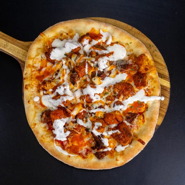 Buffalo chicken pizza with ranch drizzle on a wooden peel against a dark background.