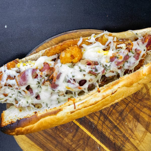 A loaded sub sandwich with melted cheese, bacon, and tater tots on a wooden board.