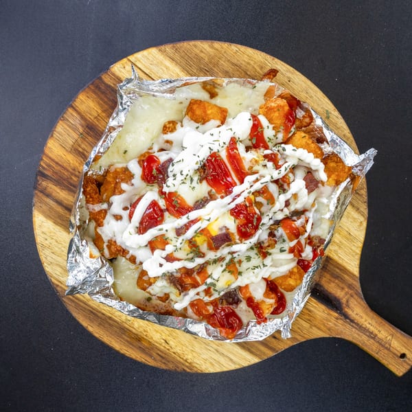 Loaded tater tots with melted cheese, ketchup, mayonnaise, and herbs on a wooden board.