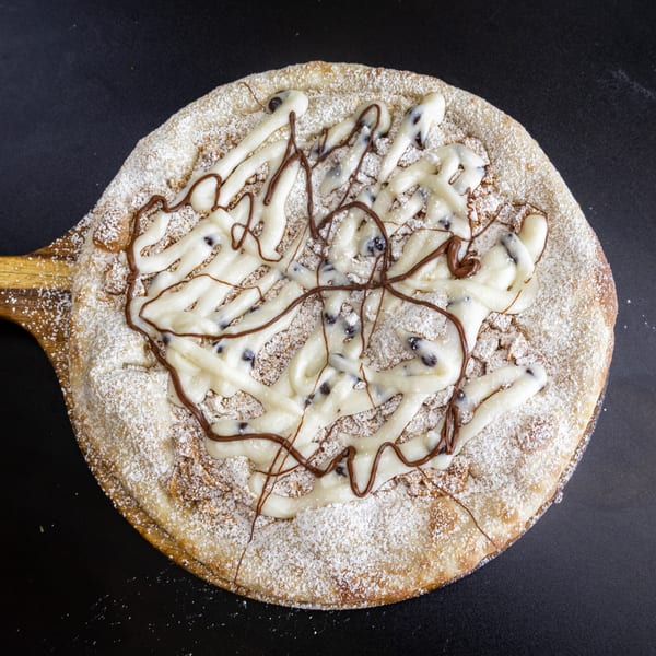 Dessert pizza on a wooden peel, topped with powdered sugar, white icing, and chocolate drizzle.