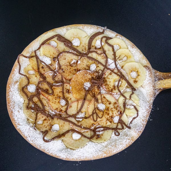Dessert pizza with sliced apples, cinnamon, powdered sugar, and chocolate drizzle on a wooden peel.